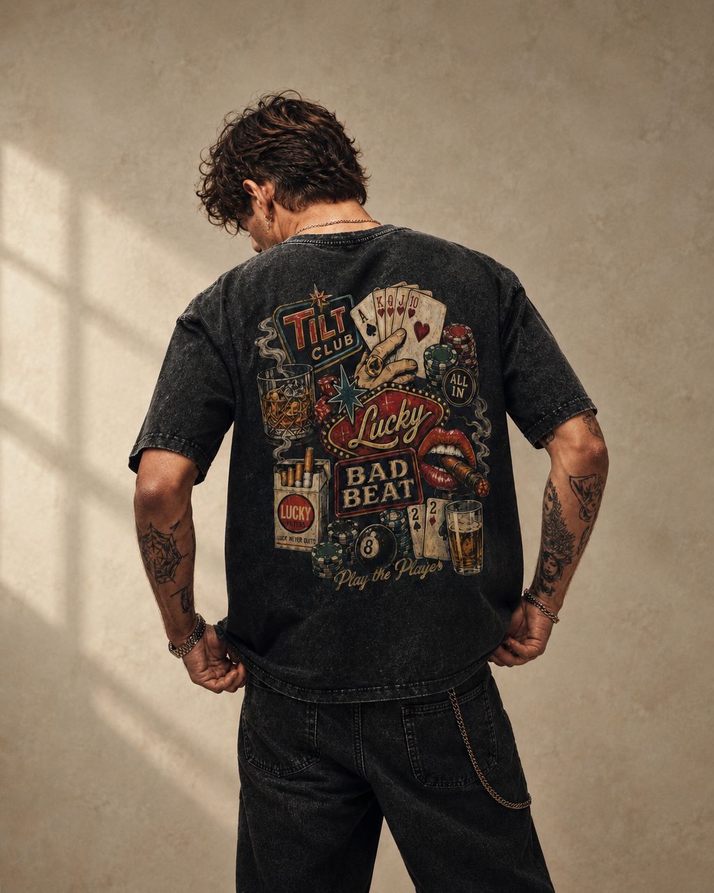 Back view of a washed black poker graphic T-shirt styled with black jeans against a warm neutral wall.